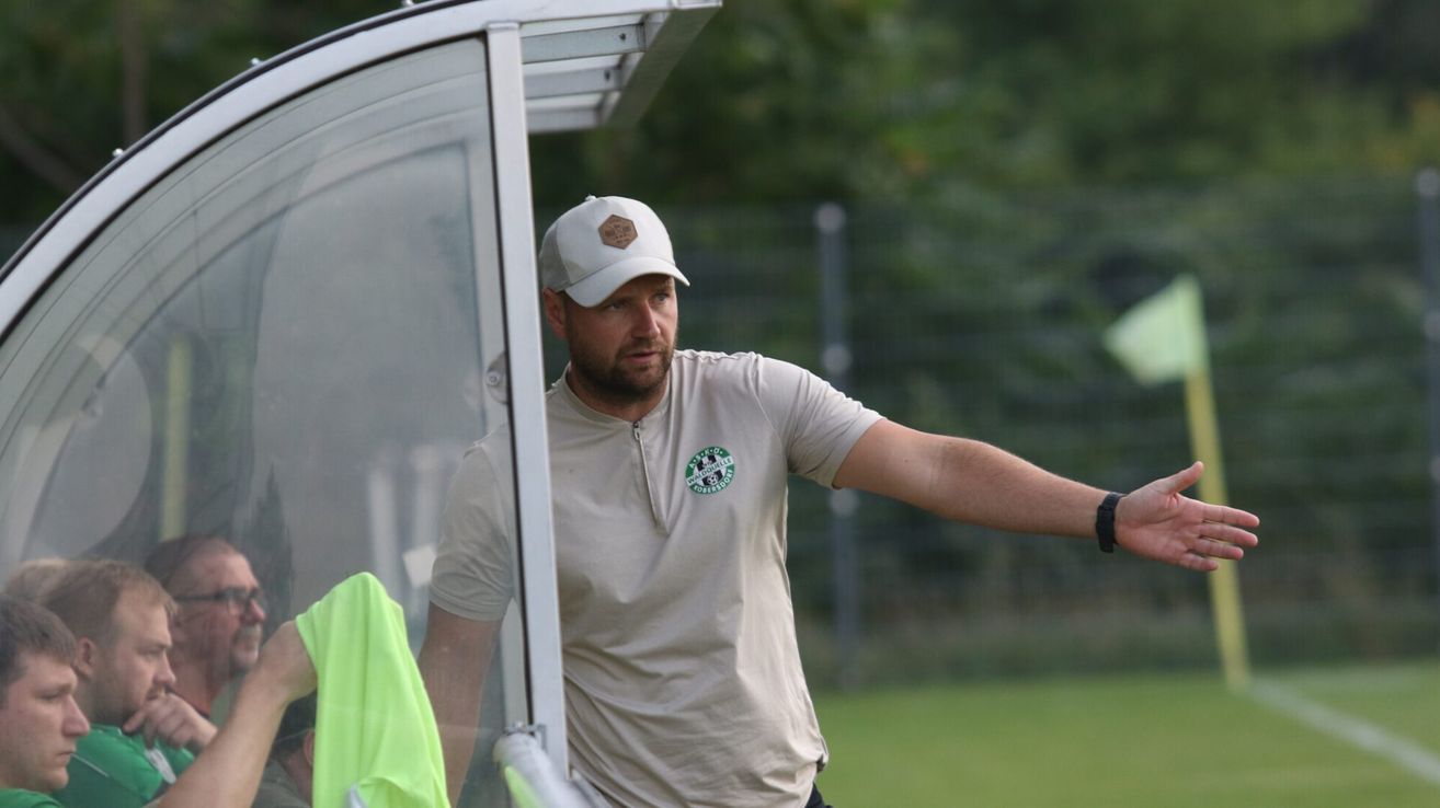 Ein Mann in einer weißen Mütze und einem beigen T-Shirt mit grünem Logo steht neben einer Glastür, möglicherweise auf einem Sportplatz.