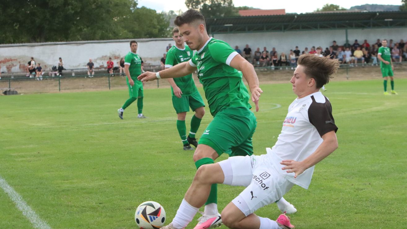 Two soccer players in green and white uniforms are competing for the ball on a grassy field. Behind them, a few players are standing, and a few people are watching from the stands.