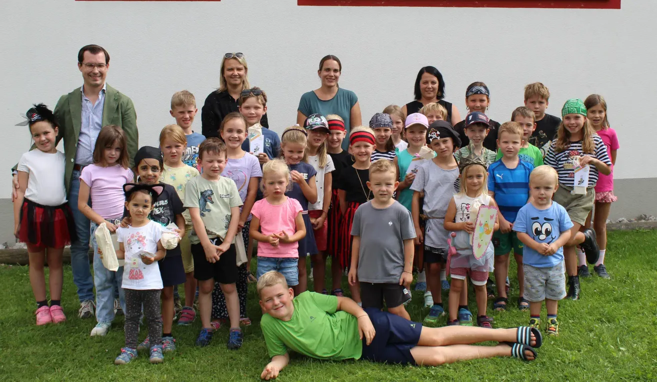 Eine Gruppe von Kindern und Erwachsenen steht und sitzt auf einem Grasfeld, einige tragen Hüte und Brillen. Sie lächeln und posieren für ein Foto.