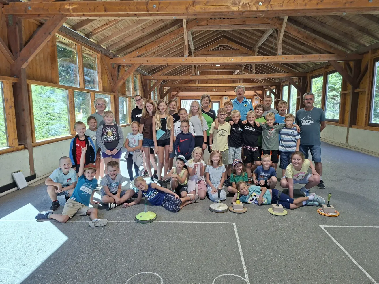 Eine Gruppe von Kindern und Erwachsenen posiert für ein Foto in einem hölzernen Raum mit weißem Boden und weißer Decke. Einige von ihnen tragen Turnschuhe.