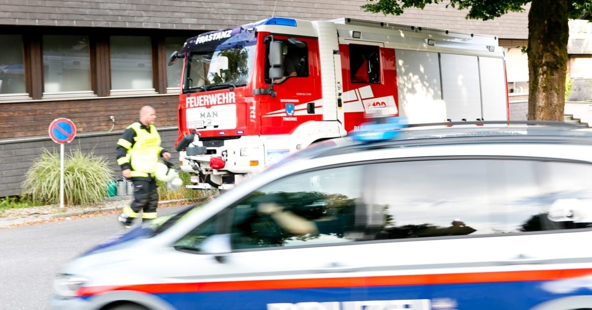 Ein Feuerwehrauto mit der Aufschrift 'Feuerwehr' steht vor einem Gebäude. Ein Polizeiauto ist in der Nähe. Eine Person in reflektierender Kleidung steht neben dem Feuerwehrauto.