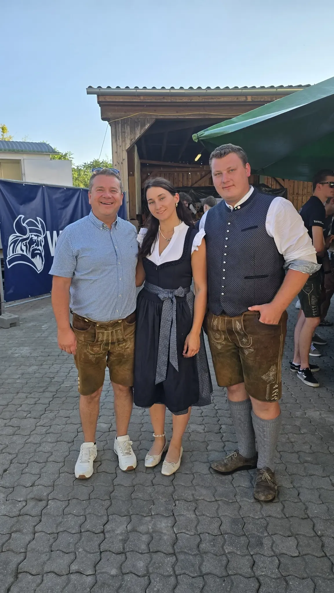 Drei Menschen in traditioneller deutscher Kleidung stehen auf einem Kopfsteinpflasterweg. Der Mann links trägt ein blaues Hemd und braune Träger, die Frau in der Mitte trägt ein schwarzes Kleid mit einer weißen Schürze und der Mann rechts trägt eine braune Weste und Träger. Hinter ihnen ist ein blauer Banner mit einem Logo zu sehen.