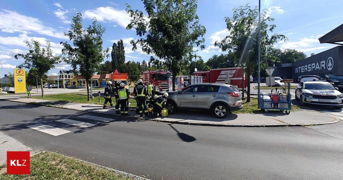 Rettungskräfte in gelben und grünen Uniformen helfen einer Person in einem silbernen Auto am Straßenrand. Bäume und Gebäude sind im Hintergrund zu sehen.