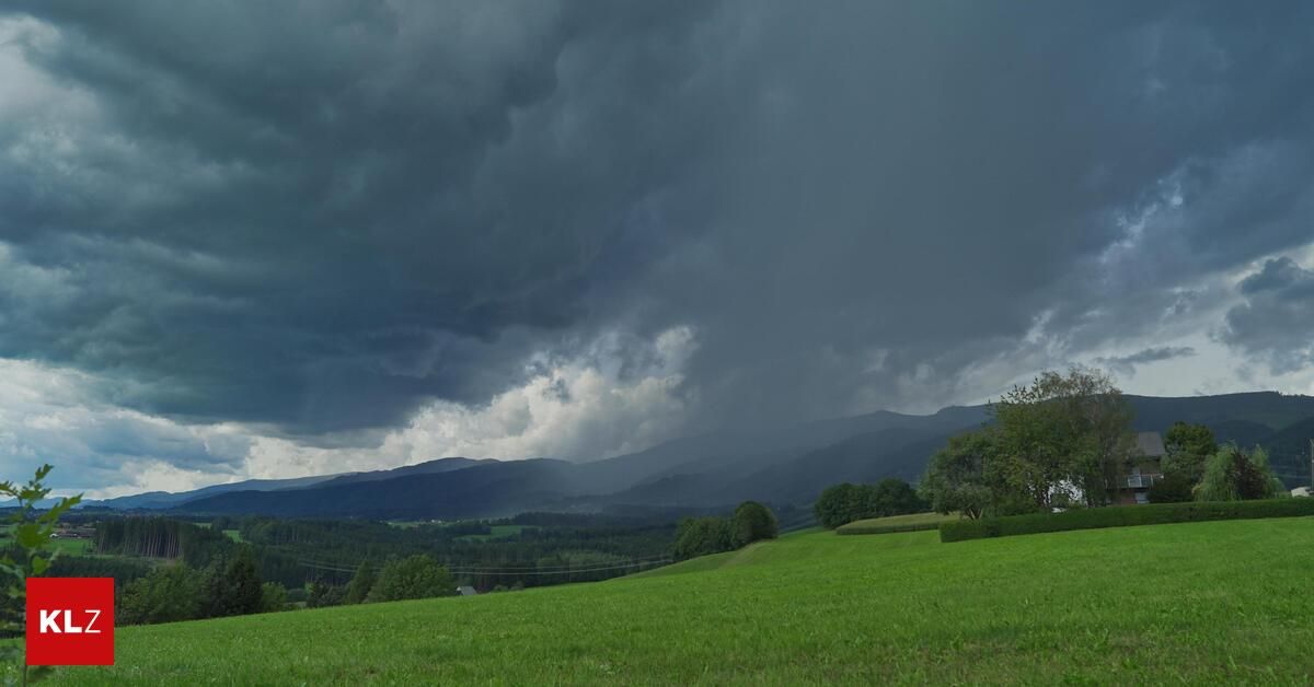 Ein grünes Feld mit einem dunklen Gewitterhimmel darüber und Bergen im Hintergrund.