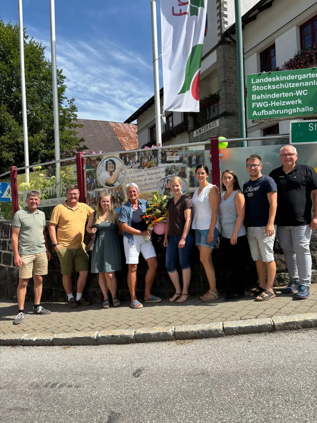 Eine Gruppe von Menschen steht vor einer Wand mit einer Collage von Fotos und einem grünen Schild mit weißem Text. Sie lächeln und posieren für ein Foto.