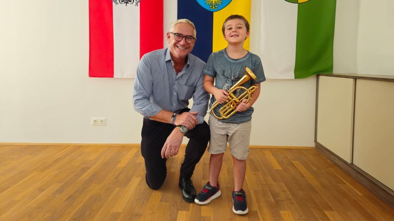 Ein Mann und ein junger Junge posieren für ein Foto in einem Raum mit Fahnen im Hintergrund. Der Junge hält eine Trompete.