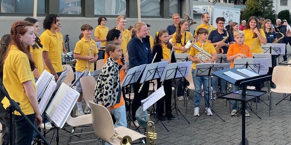 A group of children and adults in yellow shirts are gathered for a musical performance, with musical instruments and sheet music visible.