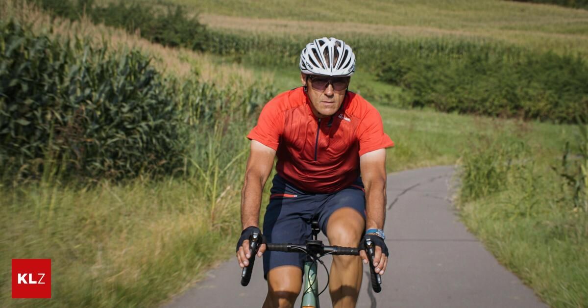 Ein Mann fährt auf einem gekrümmten Pfad durch ein Grasfeld auf einem Fahrrad. Er trägt ein rotes Shirt, blaue Shorts und einen weißen Helm.