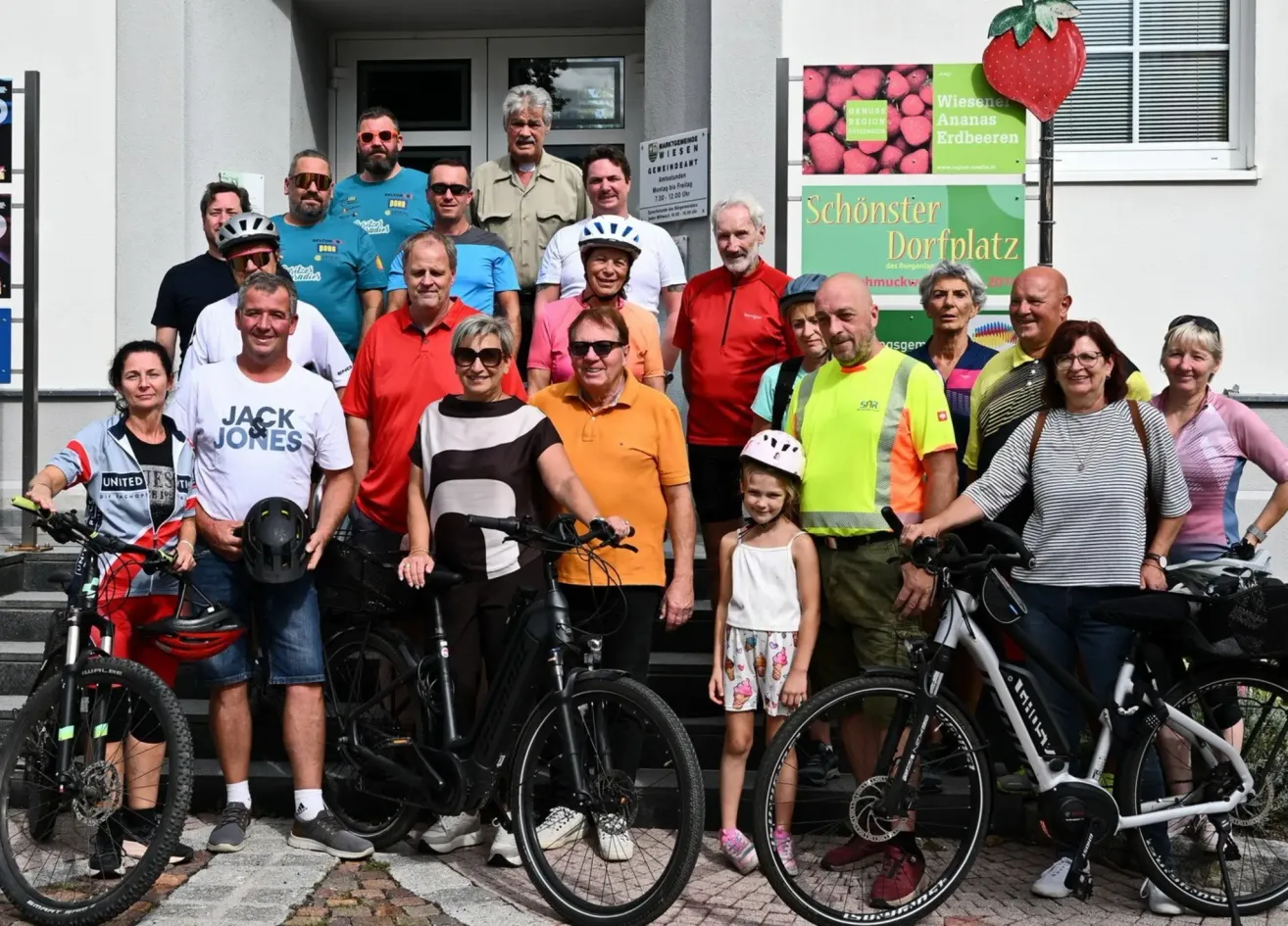 Eine Gruppe von Menschen, darunter Erwachsene und Kinder, posiert mit ihren Fahrrädern vor einem Gebäude. Einige tragen Helme und Sonnenbrillen.