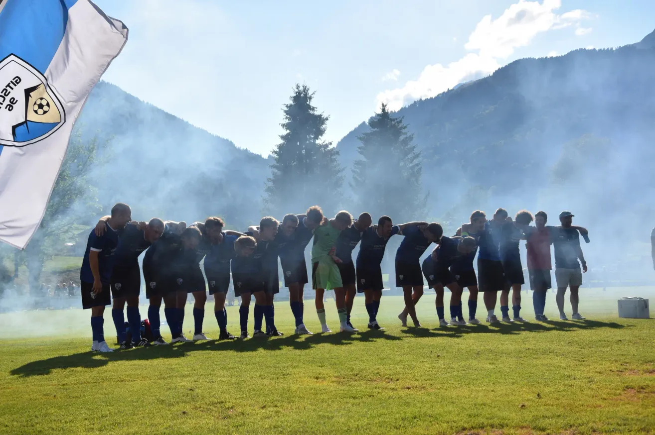 Eine Fußballmannschaft versammelt sich auf einem Rasenfeld mit Bergen und Bäumen im Hintergrund, umgeben von einer rauchigen Atmosphäre.