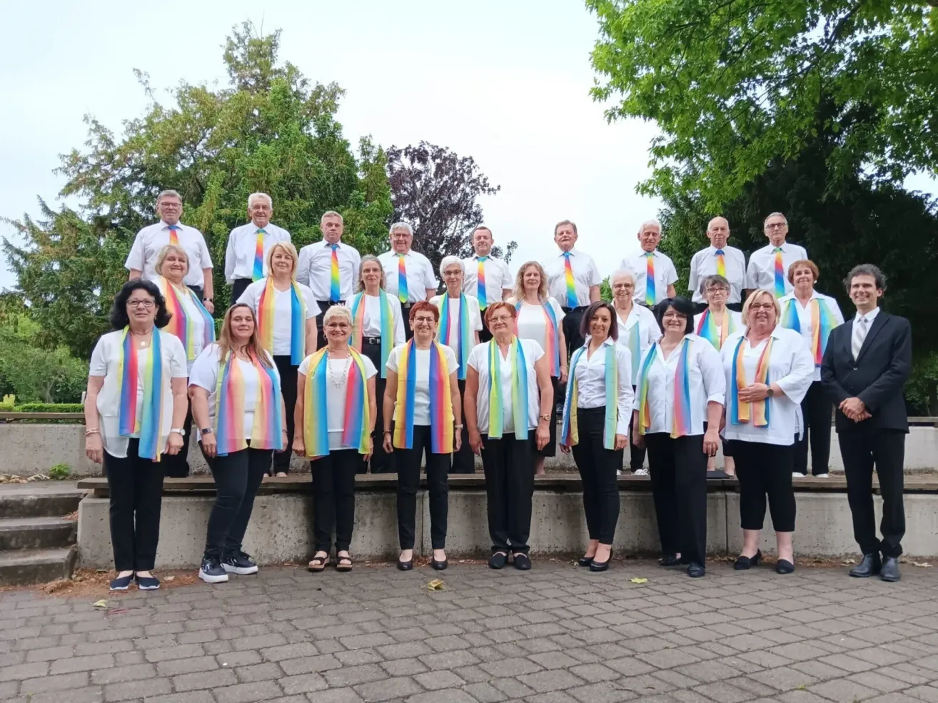A choir group stands in a row, wearing white shirts, black pants, and colorful scarves. They are in an outdoor setting with trees and a brick floor.