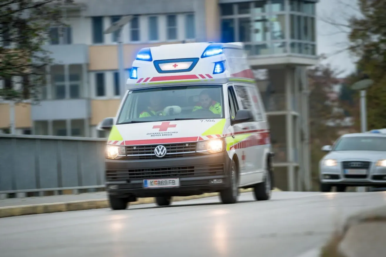 Ein Krankenwagen mit roten und weißen Markierungen fährt auf einer Stadtstraße, Lichter blinkend. Der Krankenwagen ist ein Volkswagen-Van mit einem roten Kreuzsymbol an der Seite.