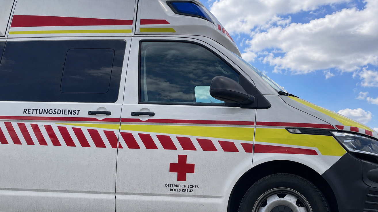 Ein weißer Krankenwagen mit roten und gelben Streifen und einem roten Kreuzsymbol an der Seite. Unter dem Kreuz steht der Schriftzug 'Rotes Kreuz'.