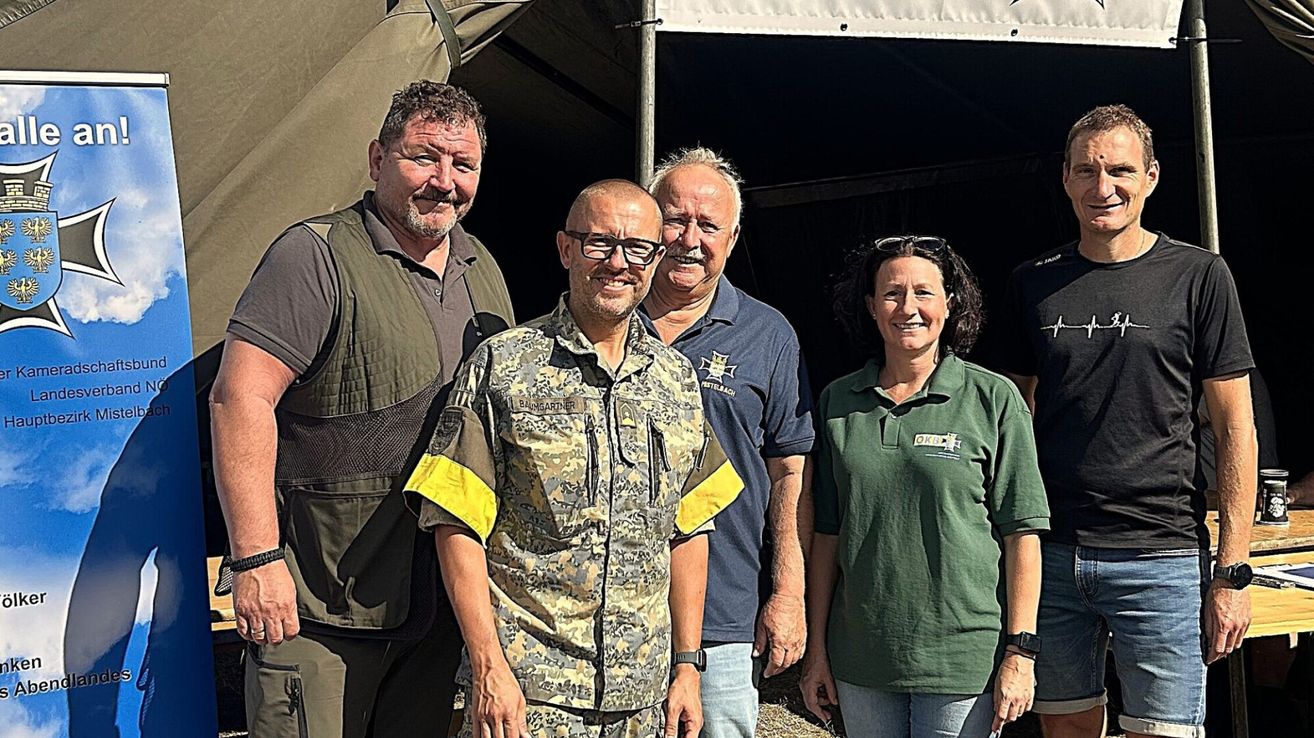 Vier Personen stehen vor einem Zelt und lächeln für ein Foto. Der Mann in der Mitte trägt eine Camouflage-Uniform mit gelben Akzenten. Der Mann auf der linken Seite hat eine Weste und eine Brille. Der Mann auf der rechten Seite trägt ein blaues Hemd und einen Schnurrbart. Die Frau auf der rechten Seite trägt ein grünes Poloshirt.