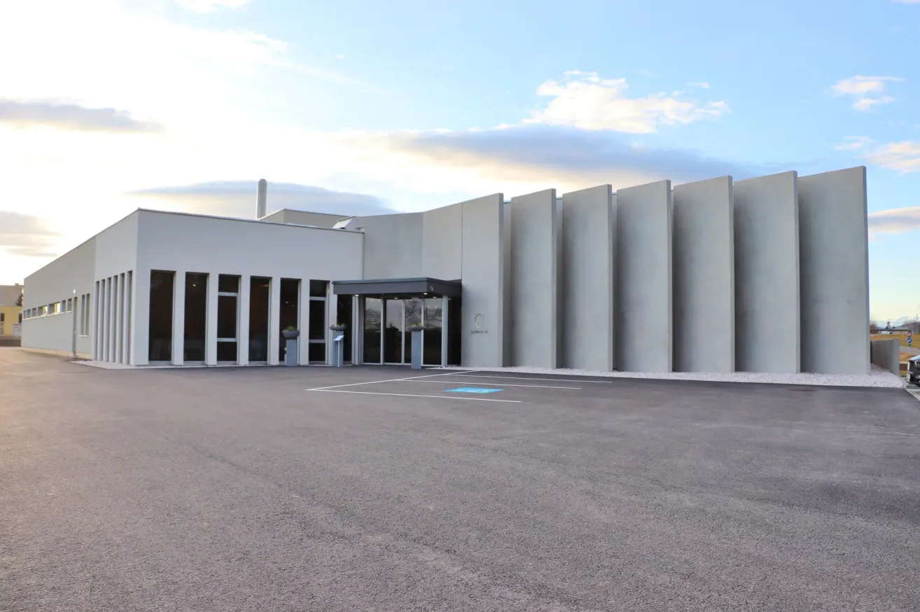 Ein modernes weißes Gebäude mit Glastüren und Fenstern steht auf einem leeren Parkplatz unter einem bewölkten Himmel.