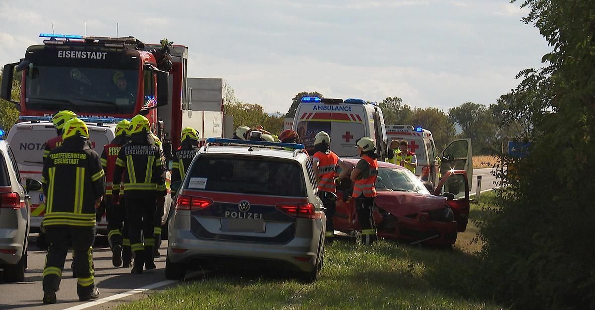 Emergency services, including firefighters and police, are attending to a car accident on the side of the road. The damaged car is red, and an ambulance is parked nearby.