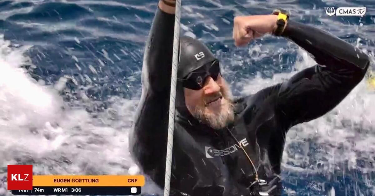 Ein Mann in einem schwarzen Neoprenanzug und Helm schwimmt im Ozean und hält sich an einem Seil fest. Er hat einen Bart und trägt eine Sonnenbrille. Das Wasser spritzt um ihn herum.