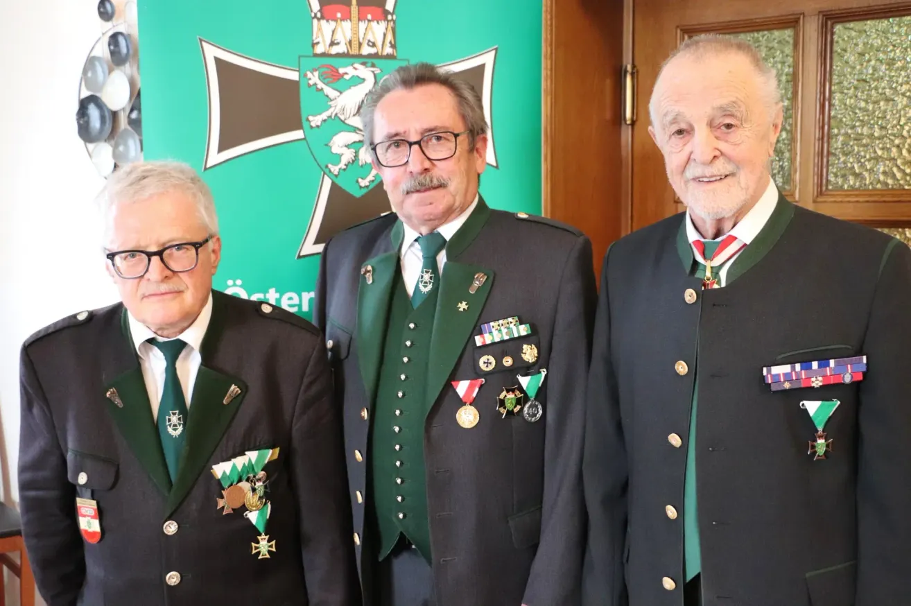 Drei ältere Männer in Militäruniformen stehen nebeneinander und lächeln für die Kamera. Sie sind mit Medaillen und Abzeichen geschmückt. Hinter ihnen ist ein grüner Hintergrund mit einem Logo.