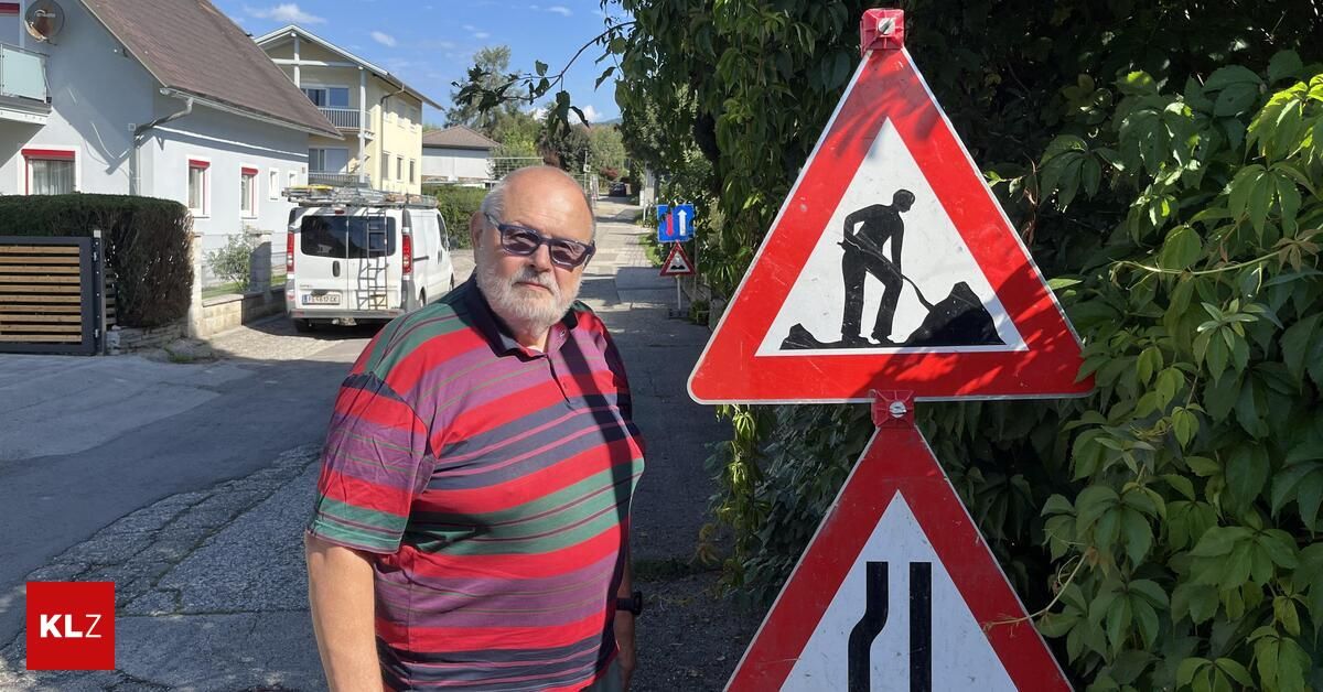 Ein älterer Mann in einem gestreiften Hemd steht neben einem Baustellenschild. Hinter ihm ist ein weißer Lieferwagen geparkt, und im Hintergrund sind Häuser zu sehen.