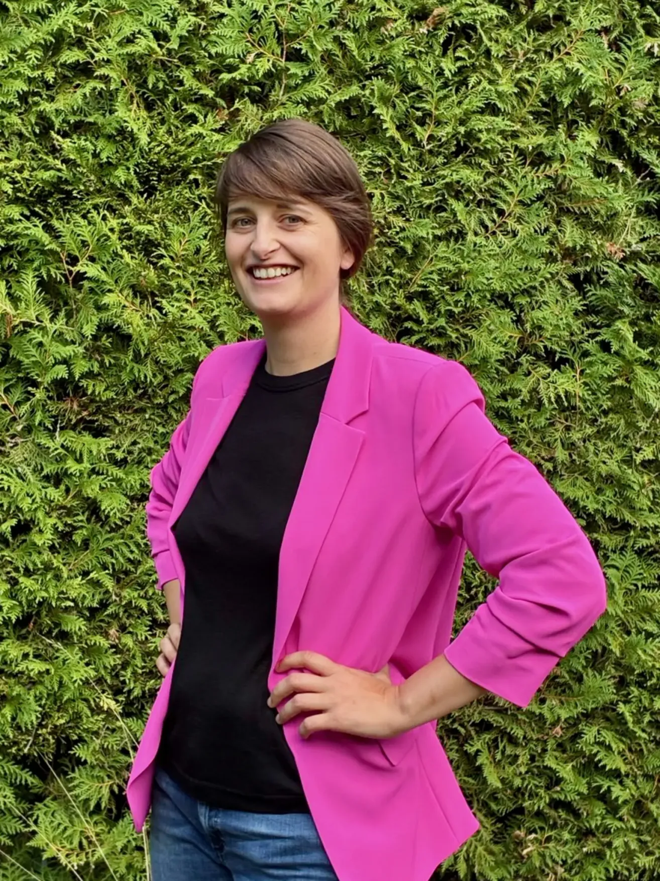 Eine Frau mit kurzen Haaren lächelt und posiert für ein Foto. Sie trägt einen rosa Blazer über einem schwarzen Oberteil. Sie hat die Hände auf der Taille und steht vor einem grünen Busch.