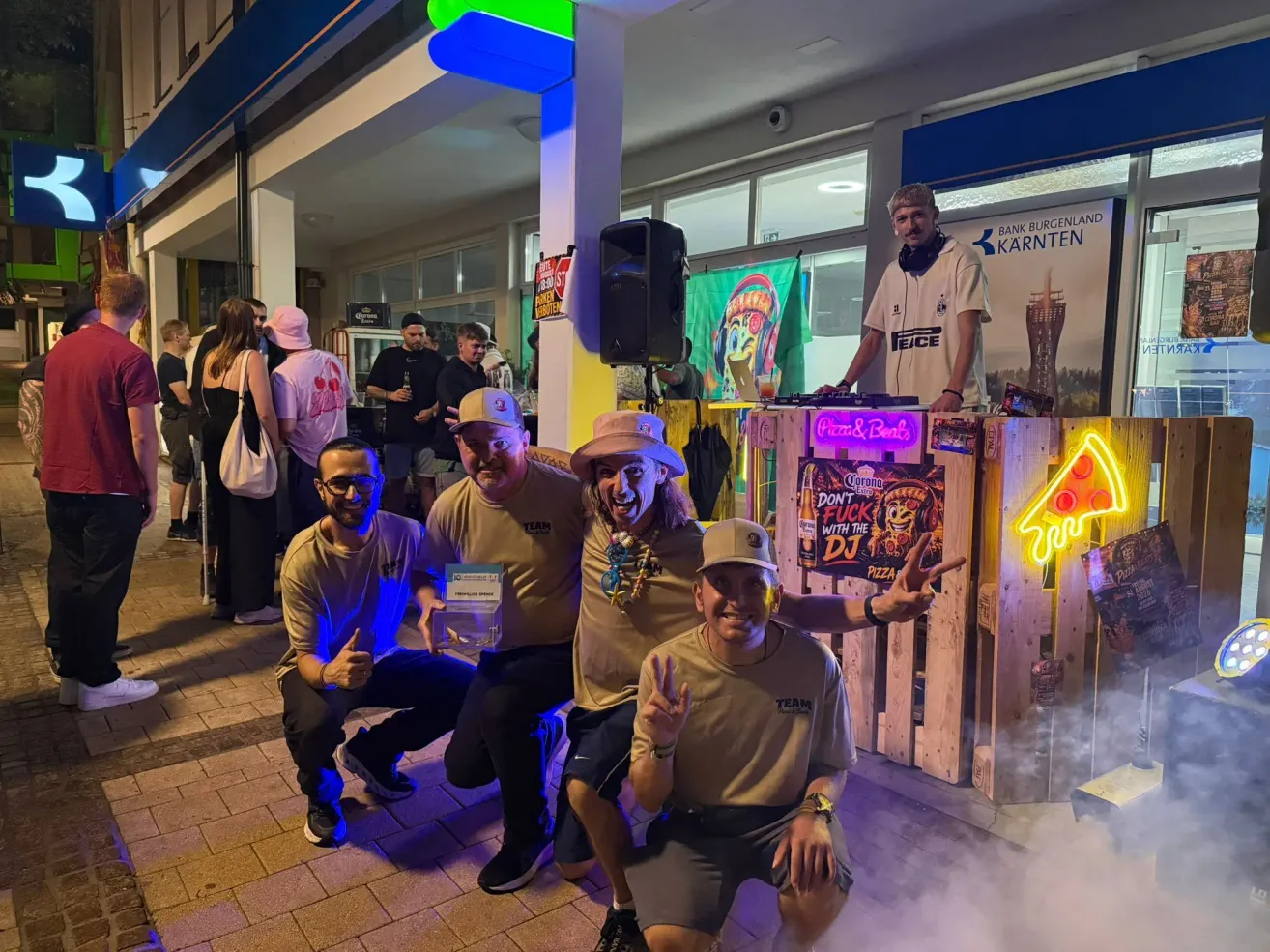 Vier Männer in passenden T-Shirts posieren für ein Foto vor einem DJ-Pult, mit einer Menschenmenge im Hintergrund. Der DJ steht hinter einem Tisch mit einem Neonschild.