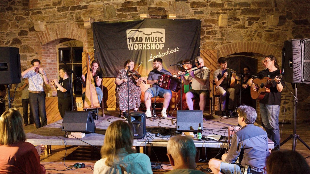 A group of musicians perform on stage at the Trad Music Workshop, with a banner in the background.