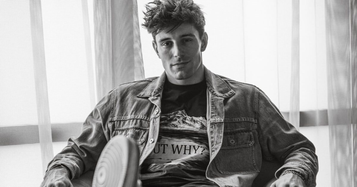 A black and white photo of a man sitting with one leg crossed, wearing a denim jacket and a t-shirt with the text 'OUT WHY?'.