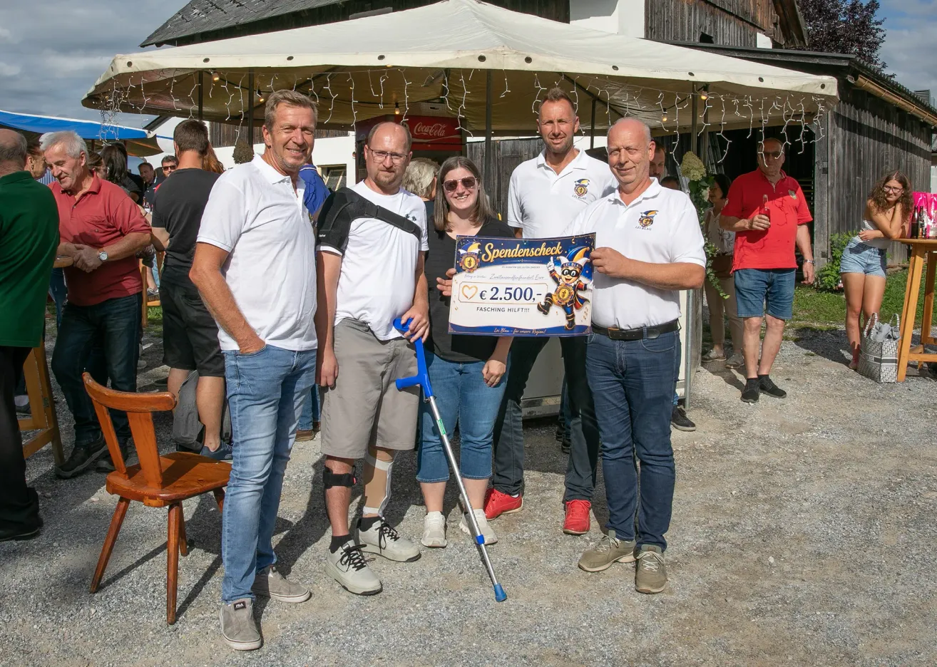 Fünf Personen stehen unter einem Zelt, lächeln und posieren für ein Foto. Eine Frau hält einen großen Scheck, und ein Mann neben ihr hat eine Krücke. Hinter ihnen stehen weitere Personen und unterhalten sich.