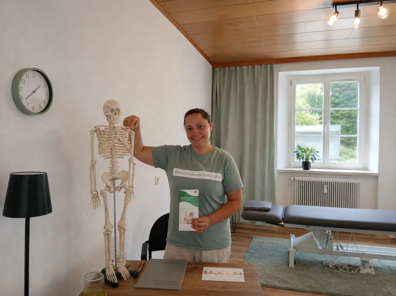 A woman holds a skeleton model while smiling and standing next to a desk with books. Behind her, a massage table and a window with a potted plant are visible.