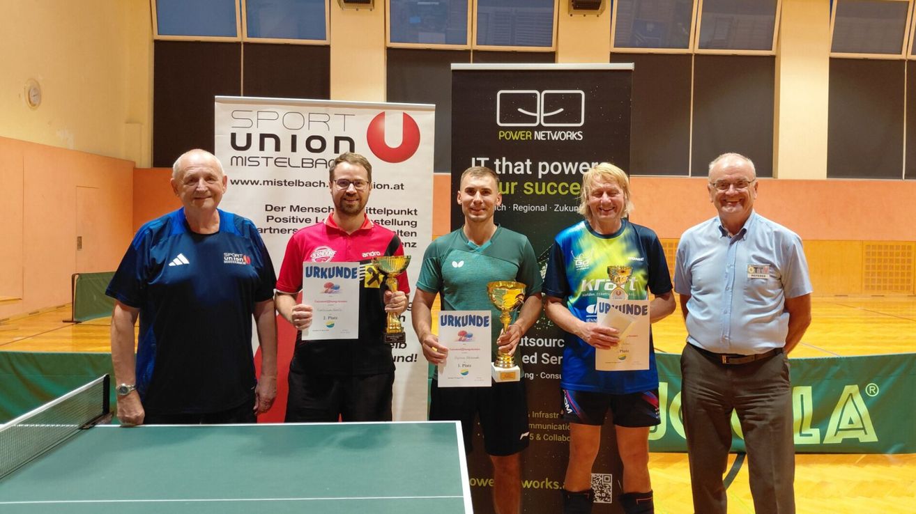 Fünf Personen stehen vor einem Banner mit einem Tischtennistisch im Vordergrund. Sie halten Pokale und Urkunden. Der Mann links trägt ein blaues Shirt, der Mann rechts ein weißes Shirt. Der Mann in der Mitte trägt ein grünes Shirt.