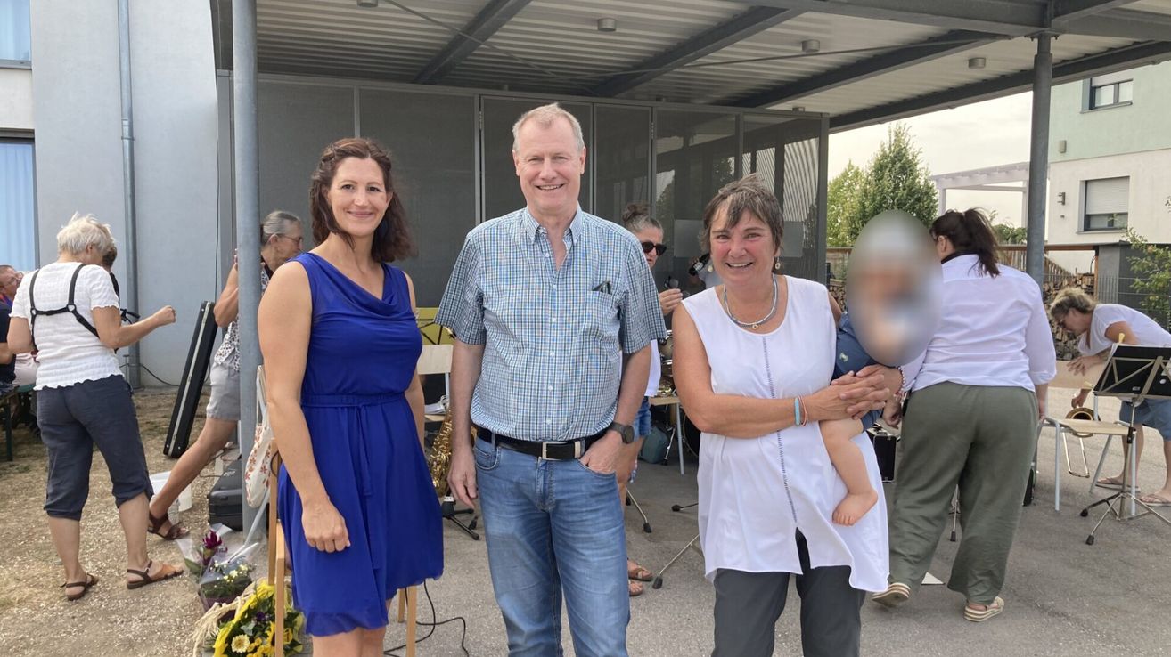 Drei Menschen lächeln für ein Foto in einem Außenbereich. Eine Frau in Blau steht links, ein Mann in einem karierten Hemd in der Mitte und eine Frau in Weiß, die ein Baby hält, rechts. Dahinter sind eine verschwommene Figur und ein Baum zu sehen.