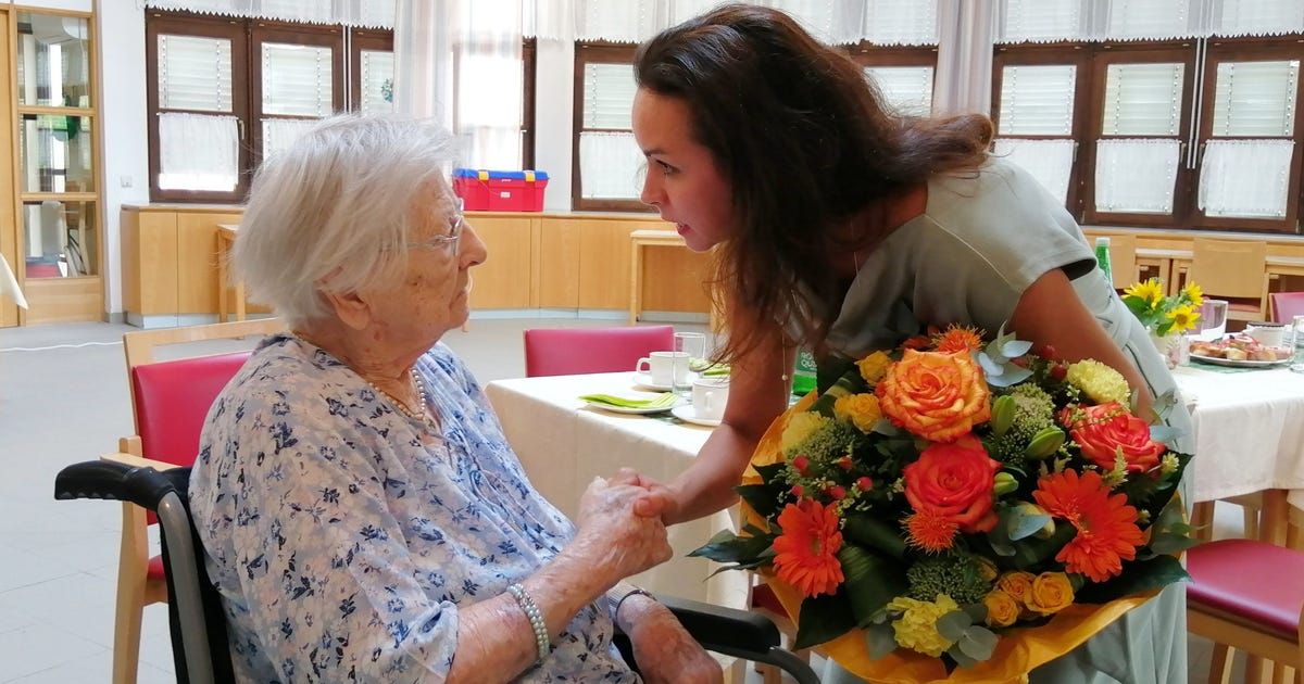 Eine ältere Frau im Rollstuhl wird von einer jungen Frau mit einem Blumenstrauß in einem Raum mit Tisch und Stühlen präsentiert.