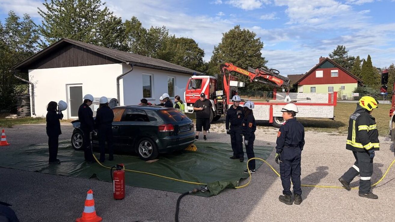 Eine Gruppe von Feuerwehrleuten in Uniform versammelt sich um ein beschädigtes Auto, einer benutzt einen Feuerlöscher. Ein Feuerwehrwagen und ein Kran sind in der Nähe.