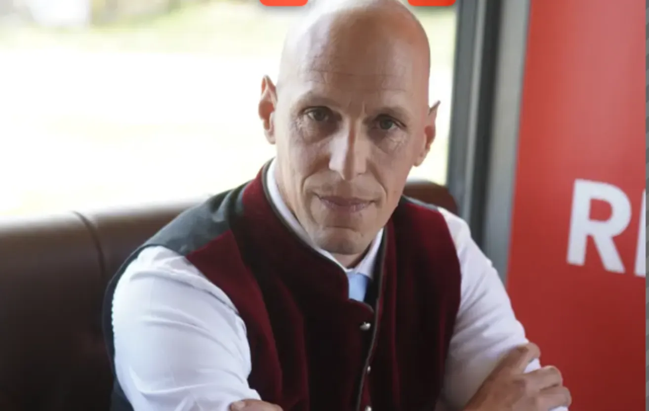 A bald man in a red vest and white shirt is smiling and looking directly at the camera.