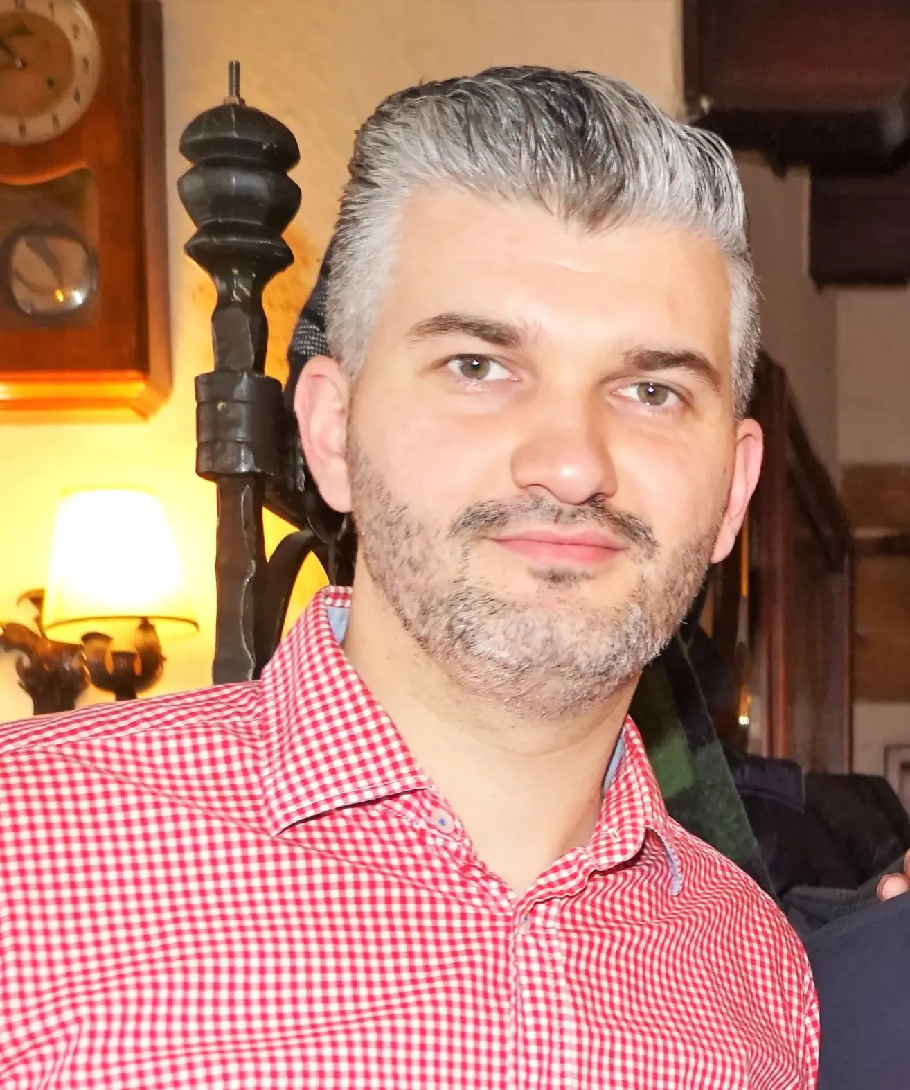Ein Mann mit grauem Haar und Bart trägt ein rot-weiß kariertes Hemd. Er steht vor einer gelben Wand.