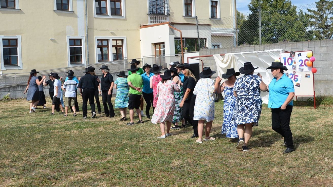 Eine Gruppe von Menschen in Hüten und Freizeitkleidung versammelt sich auf einem Rasenfeld vor einem Gebäude.