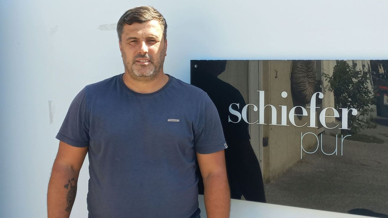 A man with a beard and mustache stands in front of a white wall with a sign that reads 'schie'. He wears a blue shirt and appears to be smiling.