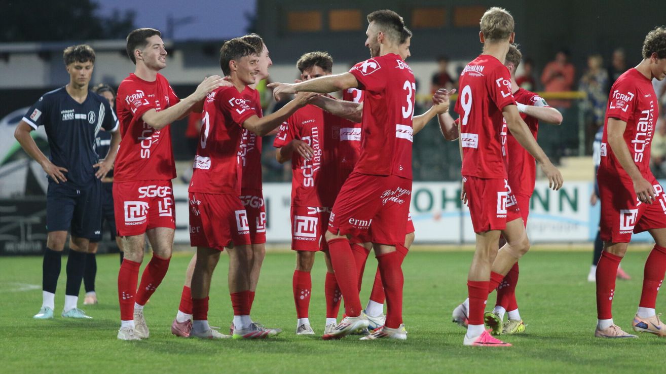 Eine Gruppe von Fußballspielern in roten Trikots feiert ein Tor auf dem Spielfeld. Sie stehen eng beieinander, einer der Spieler zeigt auf das Tor.