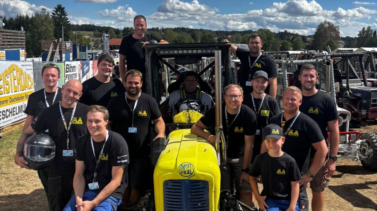 Eine Gruppe von Männern in schwarzen T-Shirts posiert für ein Foto mit einem gelben Traktor in der Mitte. Der Traktor hat eine gelbe Vorderseite und schwarze Seiten.