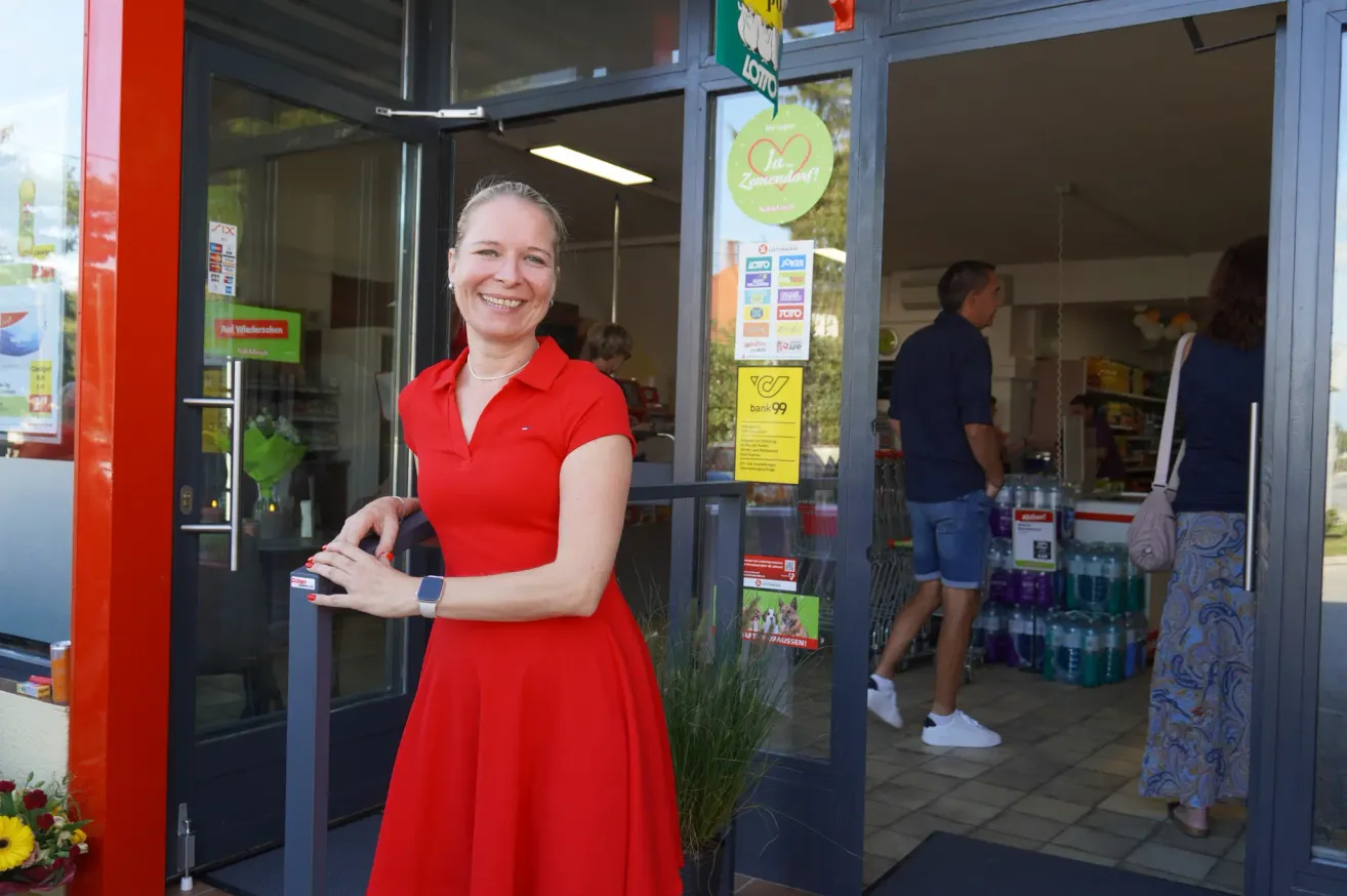 A woman in a red dress stands outside a store with a smiling face. A man in blue shorts walks past her.