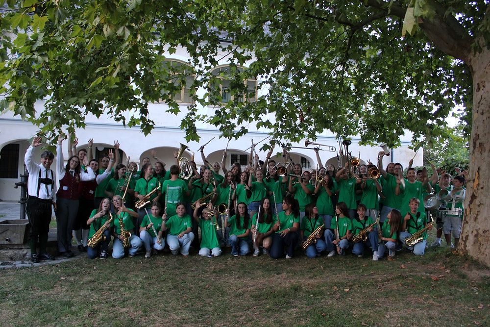 A large group of young musicians in green shirts poses for a photo under a tree in front of a building. They hold various musical instruments like saxophones, trumpets, and trombones.