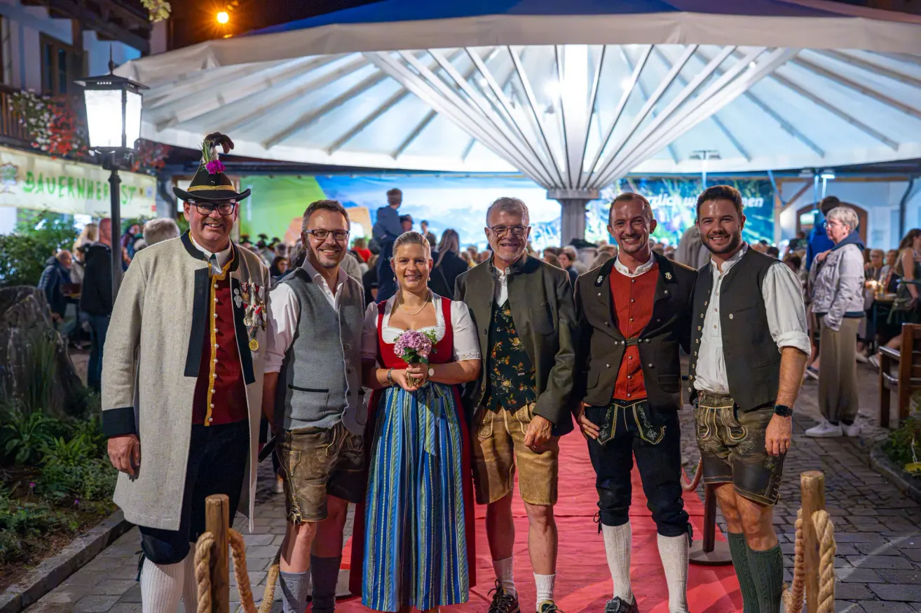 A group of people dressed in traditional Bavarian attire stand on a red carpet, smiling for a photo. The woman in the center holds a bouquet of flowers. Behind them, a large tent and a crowd of people can be seen.