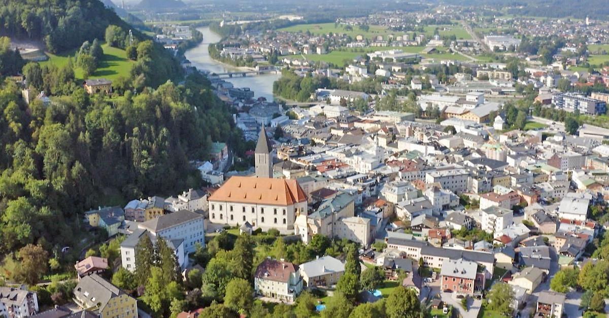 Ein Luftbild einer Stadt mit vielen Gebäuden, einem Fluss und einem grünen Feld. Die Stadt ist von Bäumen umgeben und hat ein schlossartiges Gebäude in der Mitte.