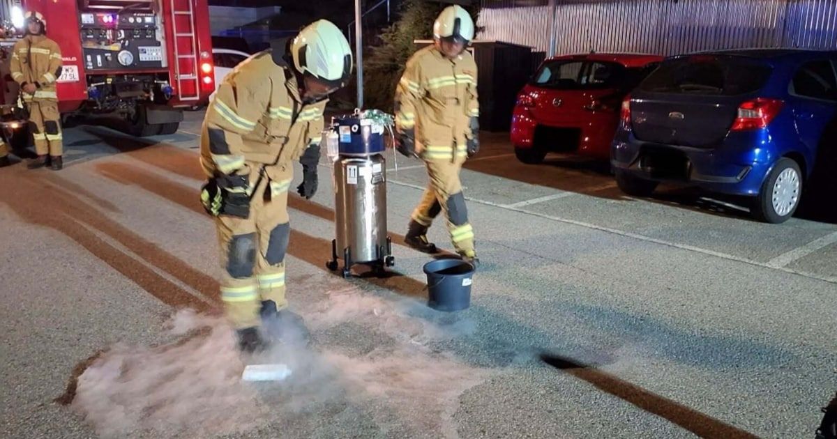 Two firefighters in full gear are cleaning a road with a bucket of water and a sprayer. A fire truck is parked nearby.