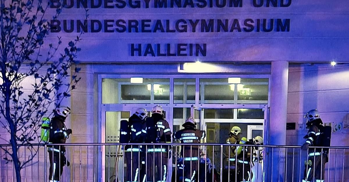 Feuerwehrleute in Schutzkleidung stehen auf einem Balkon vor einem Gebäude mit der Aufschrift 'Undesrealgynasium Hallein'. Der Eingang ist offen, und sie bereiten sich darauf vor, einzutreten. Das Gebäude ist von einem Licht beleuchtet.