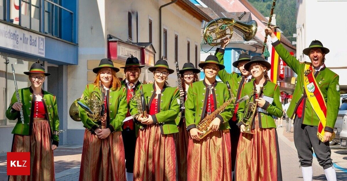 Eine Gruppe von Musikern in grünen und roten Uniformen, die verschiedene Instrumente halten, posiert für ein Foto auf einem Stadtplatz.
