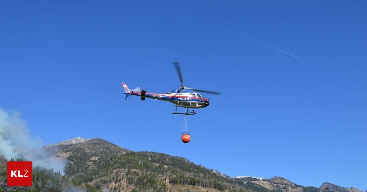 A police helicopter with the registration OE-SAX is flying in the sky, carrying a red bucket. The helicopter is flying over a mountain range with trees and a clear blue sky.