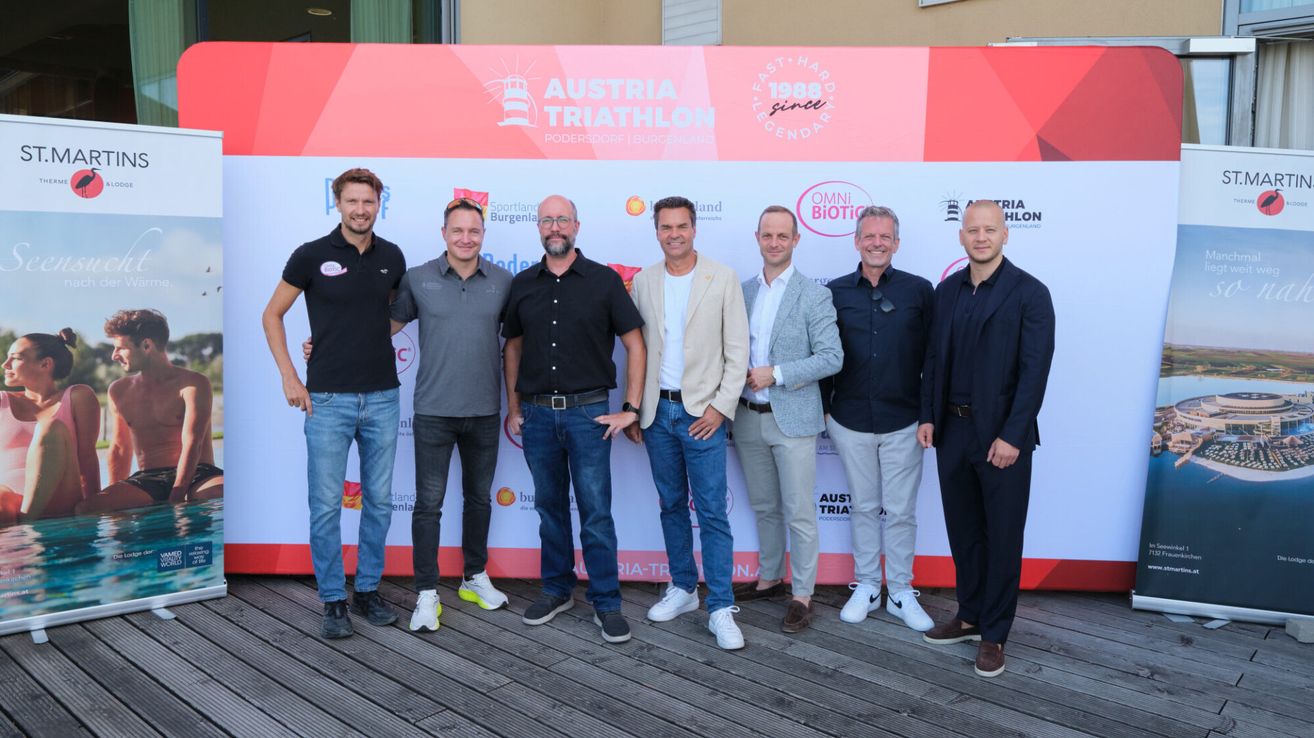 Eine Gruppe von Männern in formeller Kleidung steht vor einer Kulisse für das Austria Triathlon-Event.