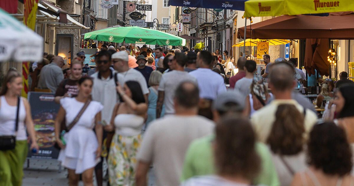 Eine geschäftige Straße voller Menschen, einige mit Hüten und Sonnenbrillen, gehen an Cafés mit Schirmen vorbei. Die Straße ist von Gebäuden und Schildern gesäumt.