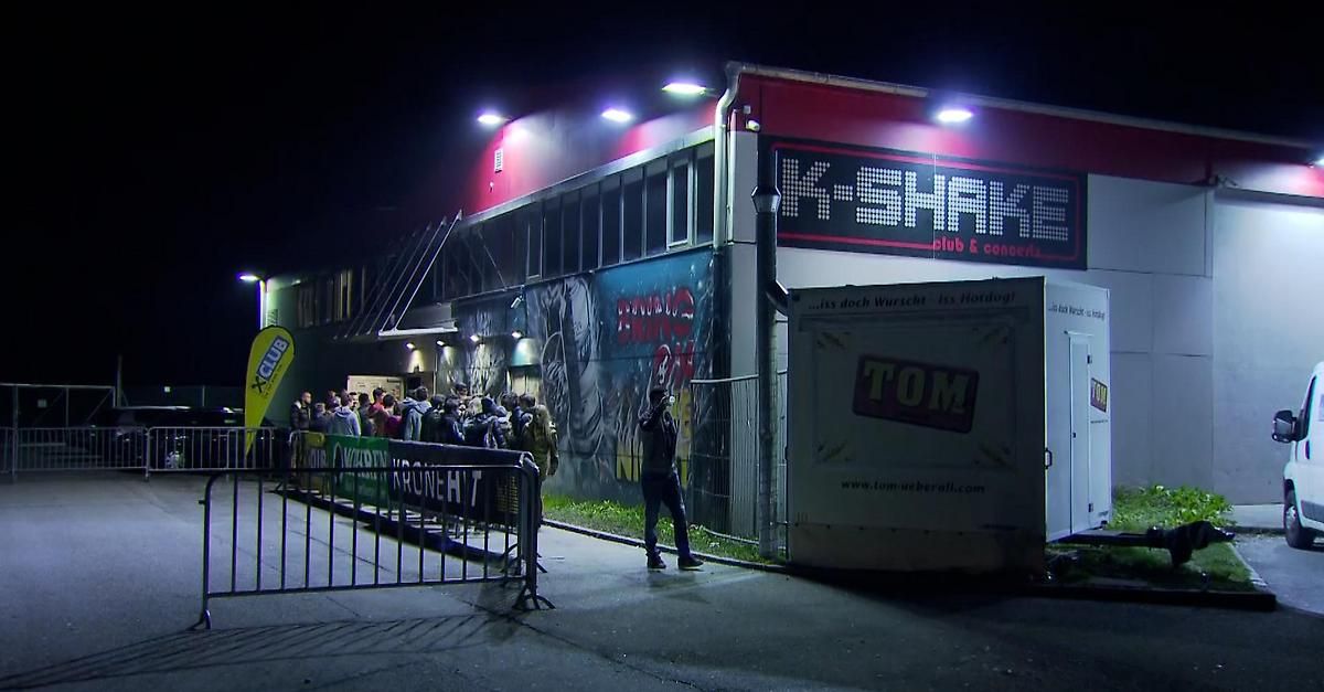 A group of people are waiting outside a building with graffiti and a sign that says K-Shake Club & Concert. A man is standing near a white container with the word Tom on it.